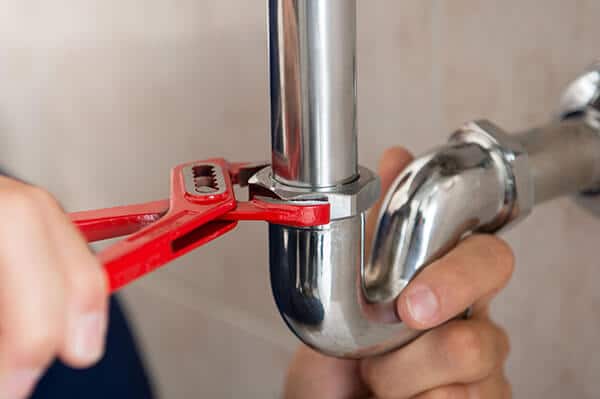 Plumber using a red adjustable wrench to tighten a pipe fitting in a residential plumbing setting, illustrating professional plumbing services related to drain maintenance and repair.