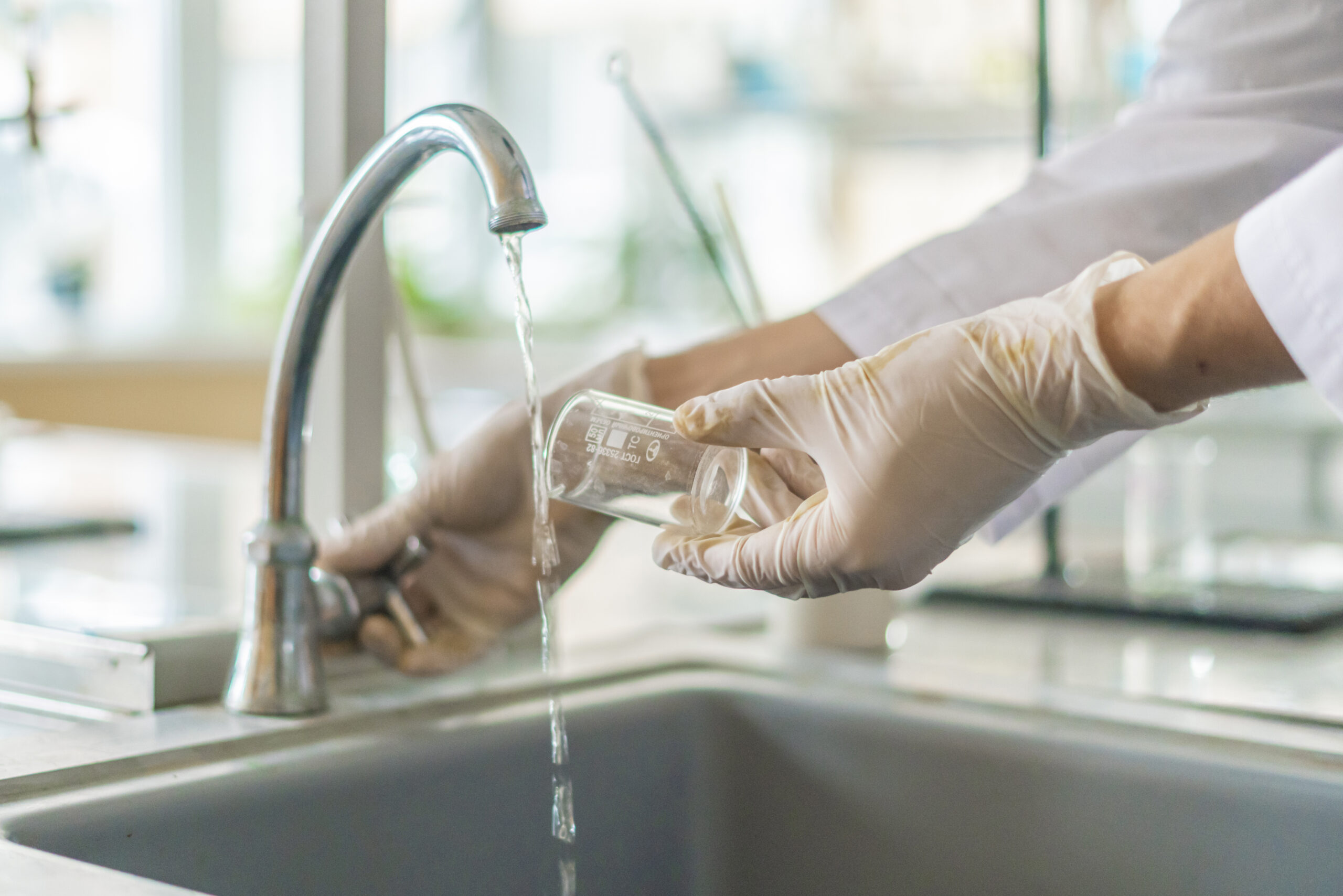 Person in gloves pouring tap water from a faucet into a glass beaker, highlighting water quality concerns in Tacoma, WA.