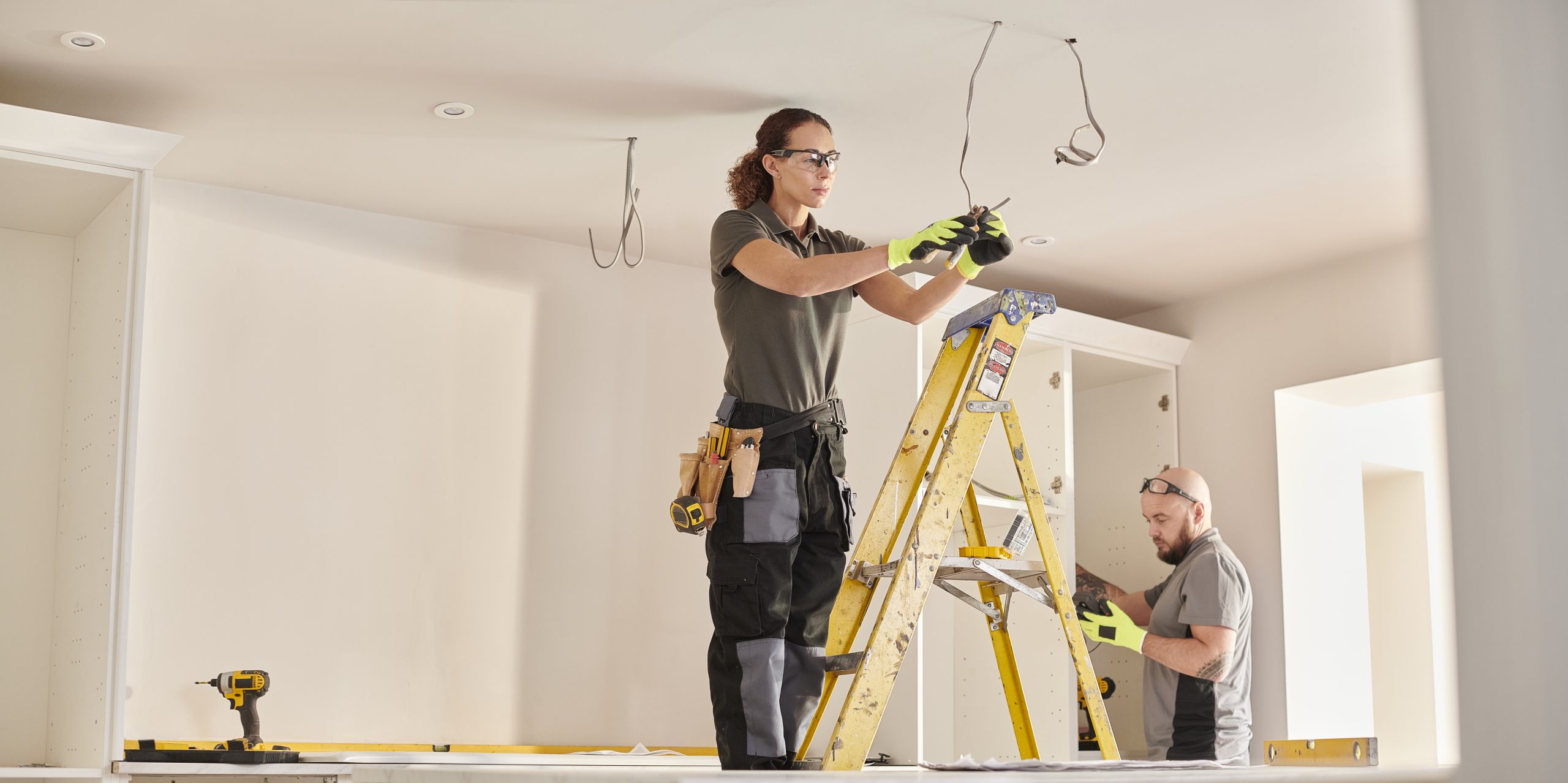 Electricians working on home electrical installation, using a ladder and tools, focused on safety and efficiency in residential setting.