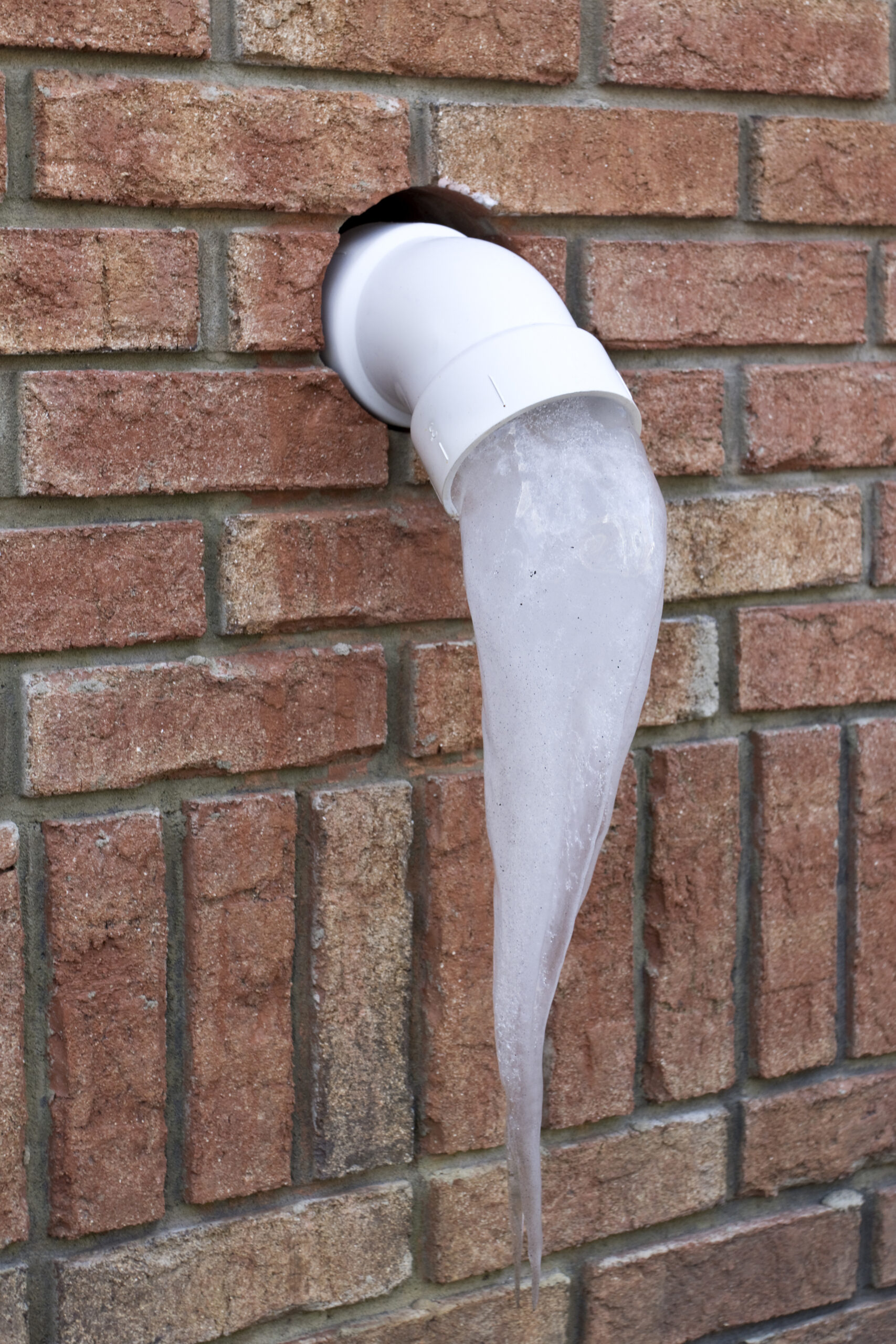 Frozen water icicle protruding from a white pipe on a brick wall, illustrating the risk of pipe freezing during winter months.