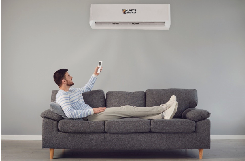 Man relaxing on a gray sofa using a remote control for a ductless air conditioning unit from Hunt's Services, showcasing modern cooling solutions for homes.